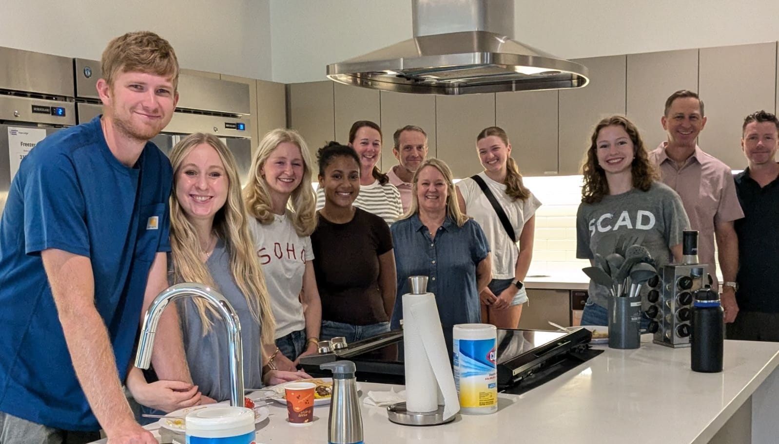 A group of smiling volunteers, including members of the PTP Gives Back team and their families, stand together in the kitchen of Hope Lodge Dallas after preparing and serving a meal for the residents.