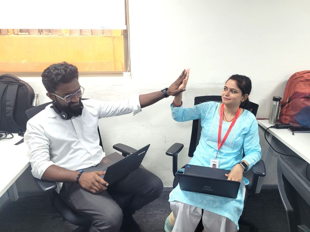 Two employees smiling and giving a high five while working on laptops in an office setting.