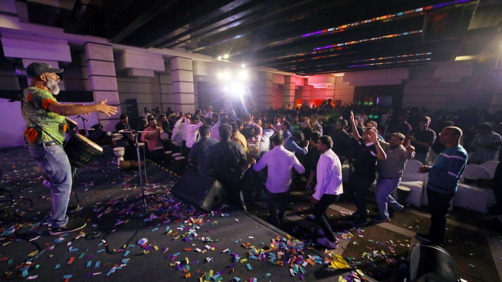 Crowd of Stratogent, now a PTP company employees dancing and celebrating indoors with confetti on the floor while a performer plays drums on stage during a Stratogent company event.